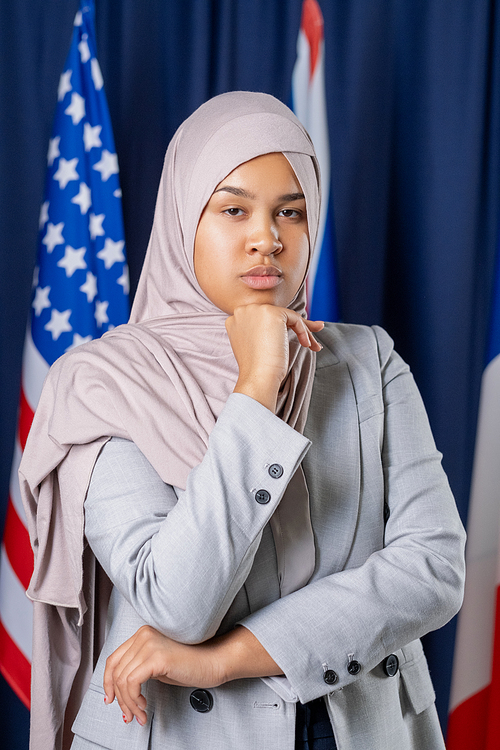 Young serious female politician in hijab and formalwear looking at you while standing against flags of several countries