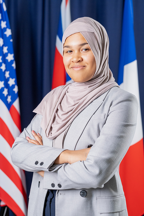 Young well-dressed Muslim female politician in hijab and elegant suit standing in front of camera on background of several flags