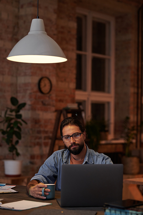 Bearded businessman drinking coffee and looking at monitor of laptop while working at the table at office