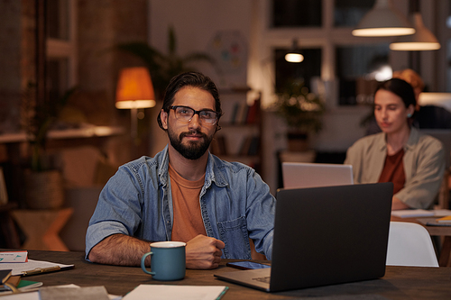 Portrait of businessman looking at camera while sitting at his workplace with laptop and working at office with his colleagues in the background