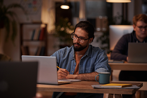 Young bearded businessman in eyeglasses making notes and working on laptop at the table at office