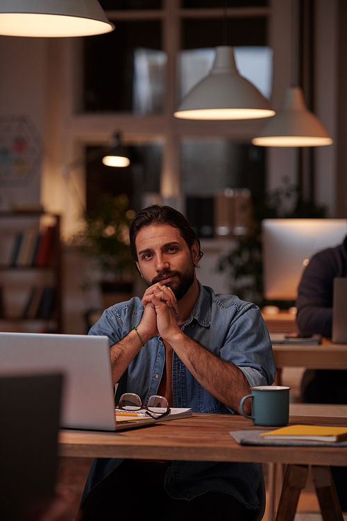 Portrait of young designer in casual clothing sitting at his workplace in front of laptop and looking at camera