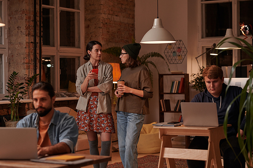 Two young women talking together while standing at office with their colleagues working at their workplaces