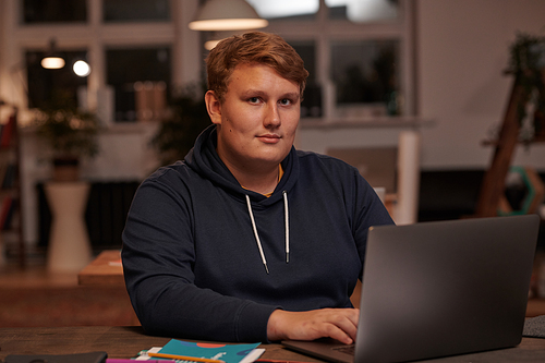 Portrait of young businessman looking at camera while sitting at the table working online on laptop at office