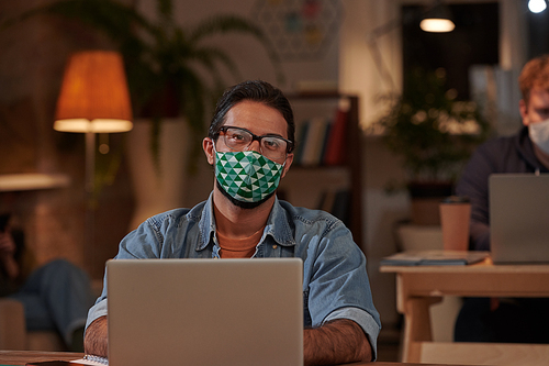 Portrait of graphic designer in protective mask looking at camera while working on laptop at the table at office