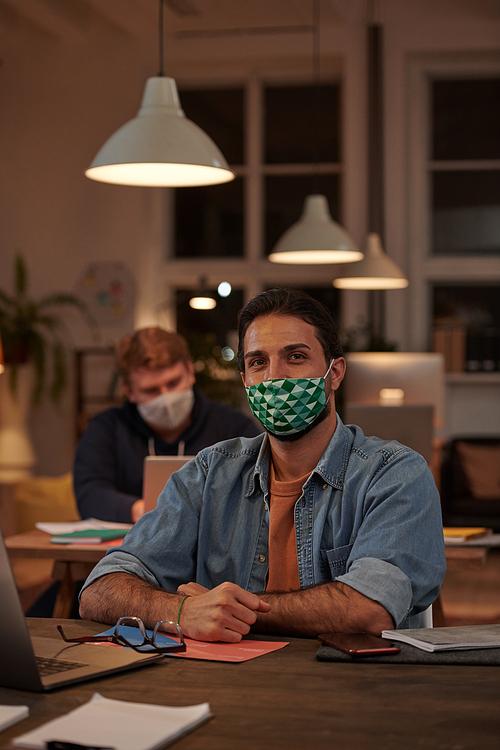 Portrait of young businessman in protective mask looking at camera while working at the table at office in the evening