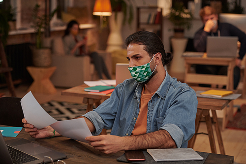 Young businessman in casual clothing working in protective mask at his table with documents at office