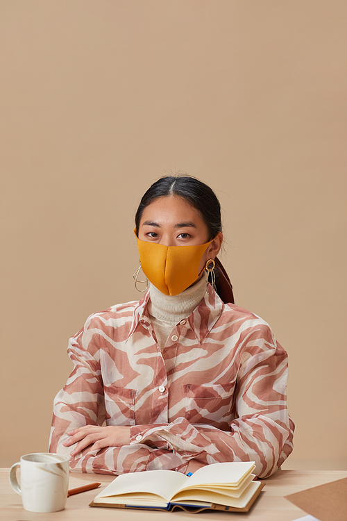 Portrait of Asian woman in protective mask looking at camera while sitting at the table and studying