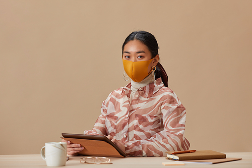 Portrait of Asian woman in protective mask looking at camera while sitting at the table and using digital tablet