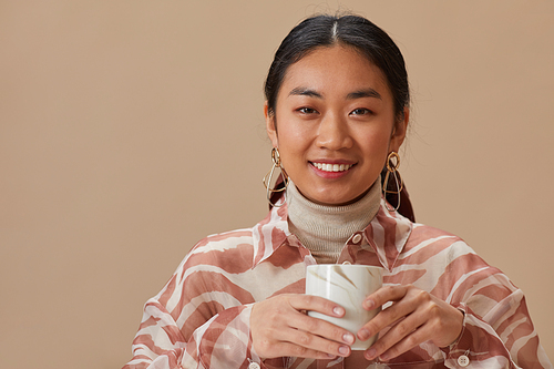 Portrait of Asian young woman smiling at camera holding mug with tea isolated on beige background