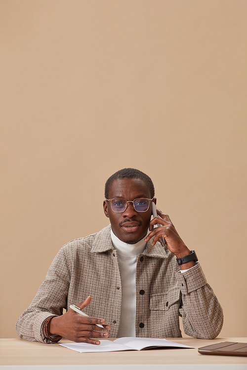 Portrait of African man sitting at the table looking at camera while talking on mobile phone isolated on beige background