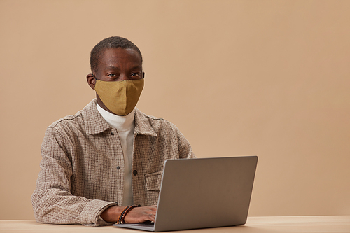 African man in protective mask sitting at the table and typing on laptop computer isolated on beige background