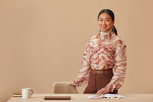 Portrait of Asian young businesswoman smiling at camera standing near her workplace against the beige background