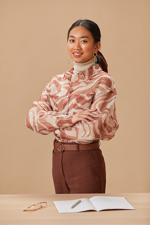 Portrait of Asian young teacher standing with arms crossed near the table and smiling at camera