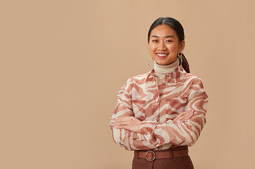 Portrait of Asian beautiful woman in elegant clothing standing with arms crossed and smiling at camera against the beige background