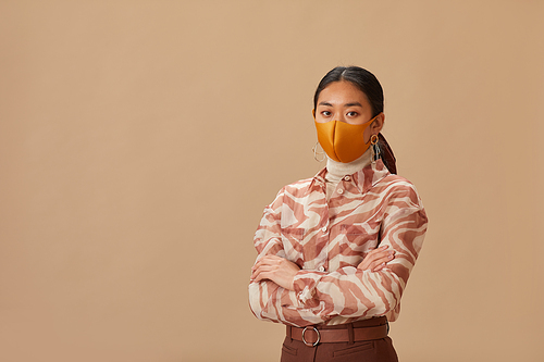 Portrait of Asian young woman in protective mask standing with arms crossed looking at camera against the beige background