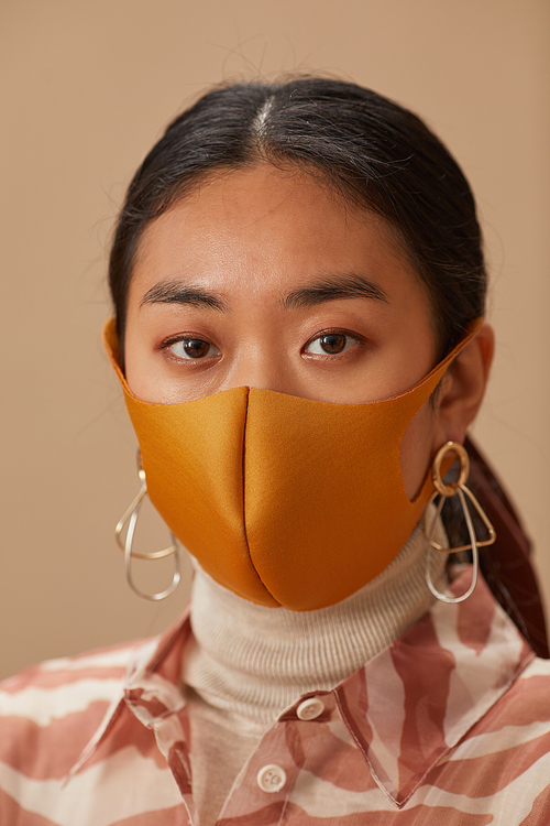 Close-up of Asian woman looking at camera wearing protective mask against the beige background