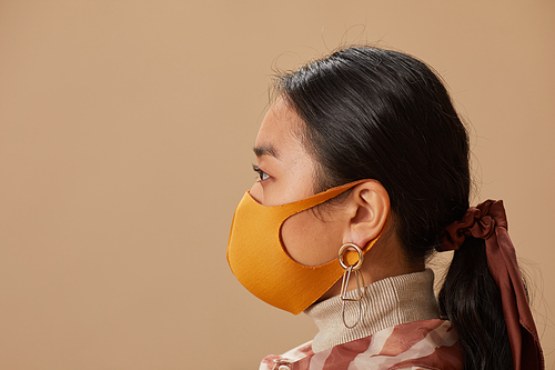 Side view of Asian young woman wearing protective mask standing against the beige background