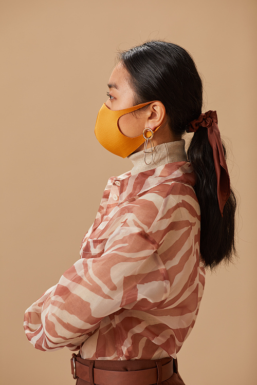 Side view of Asian young woman in elegant clothes wearing protective mask isolated on beige background