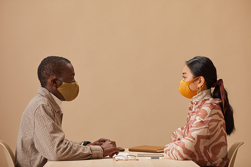 Side view of two colleagues in protective masks sitting at the table and looking at each other during meeting