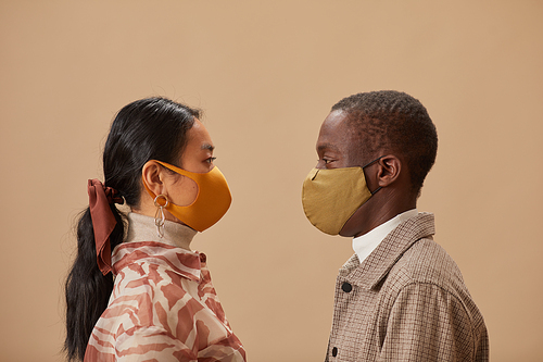 Side view of multiethnic young couple in protective masks looking at each other isolated on beige background