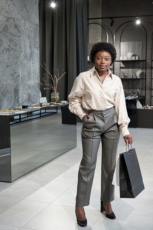 Young African female consumer with paperbags standing against display with jewelry assortment in boutique