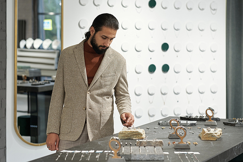 Young elegant man standing by table with luxurious jewelry while choosing accessory for himself or his girlfriend