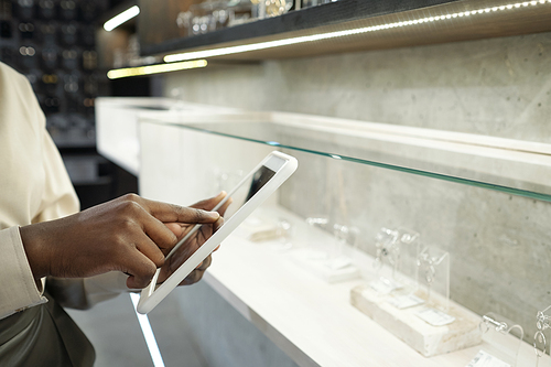 Hand of young contemporary shop assistant pointing at tablet screen while standing against large display with jewelry collection