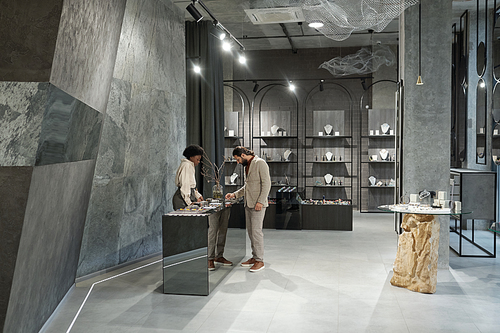 Interior of jewelry boutique with male buyer and shop assistant standing by display with assortment of necklaces, bracelets and earrings