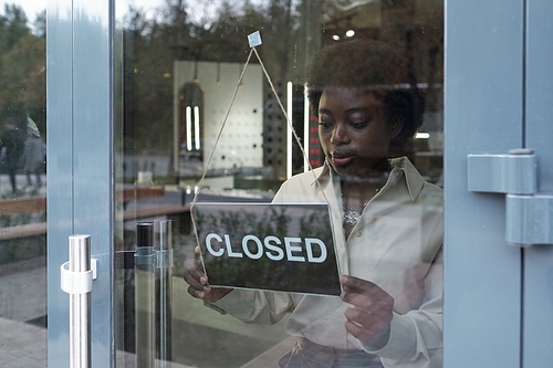 Young African shop assistant hanging closed sign on door of luxurious jewelry boutique at the end of working day