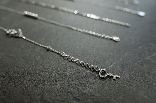 Silver necklace or bracelet with tiny decorative key over dark grey background against other jewelry items