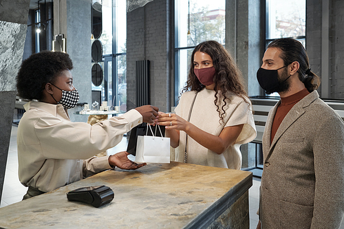 Young woman in protective mask taking paperbags with purchase held by African female shop assistant over counter in jewelry store