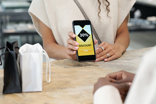 Hands of young consumer showing discount coupon in smartphone to shop assistant before paying for purchase