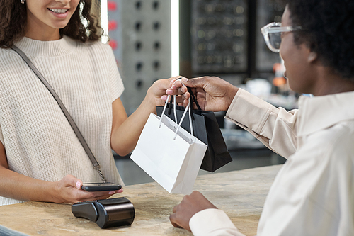 Shop assistant passing paperbags with bought jewelry to happy young female consumer paying for purchase by counter