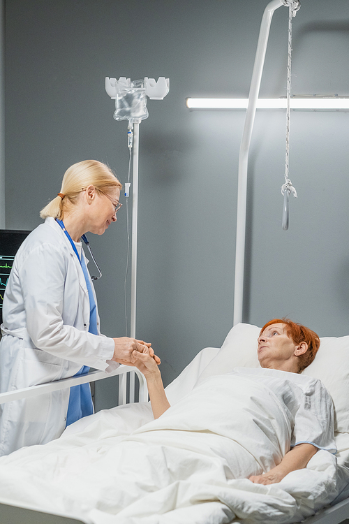 Mature female doctor holding hand of her patient and talking to her while she lying on hospital bed