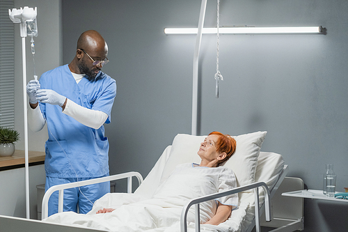 African anesthesiologist in uniform making drip for senior woman before operation in hospital ward