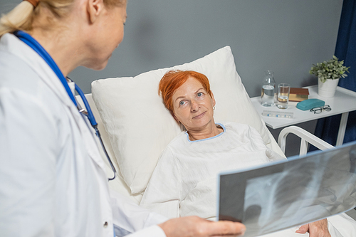 Mature woman lying on the bed and listening to the doctor who speaking about the x-ray image in the ward