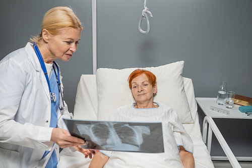 Mature doctor in white lab coat showing the x-ray image to elderly woman who lying in bed at hospital ward