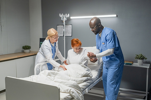 Doctor with nurse helping to stand the senior woman from the bed in the hospital ward