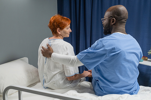 Rear view of African male doctor in uniform embracing and talking to mature woman while they sitting on the bed in the ward