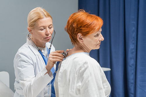 Female doctor in white coat checking the heart of senior woman with stethoscope while they sitting in the hospital ward