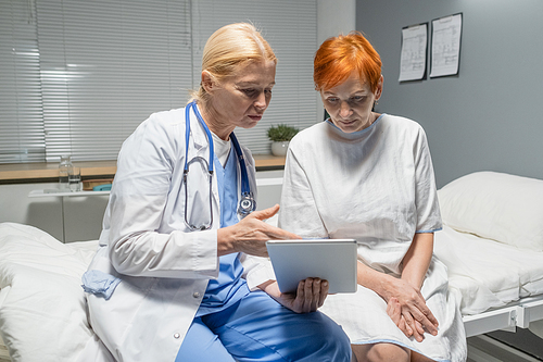 Mature female doctor in white coat pointing at digital tablet and discussing treatment together with senior patient in hospital ward