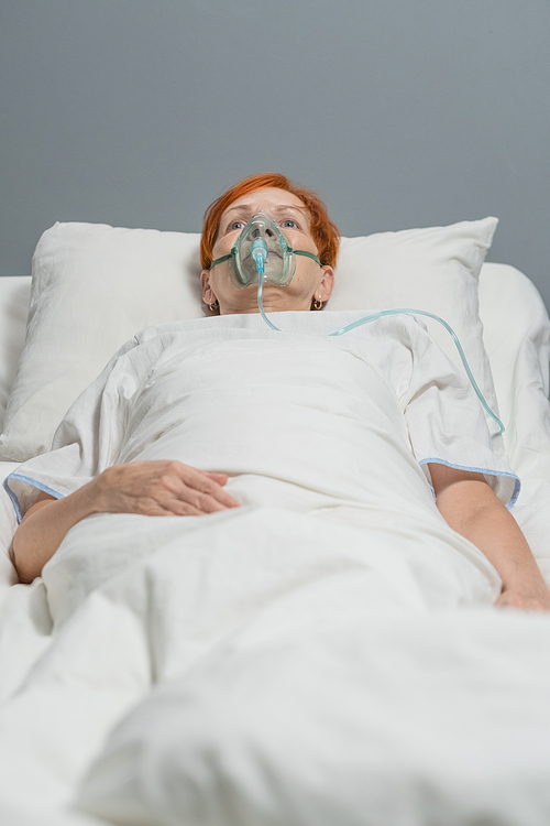 Senior woman in oxygen mask lying on the bed and breathing with special medical equipment in hospital ward during coronavirus