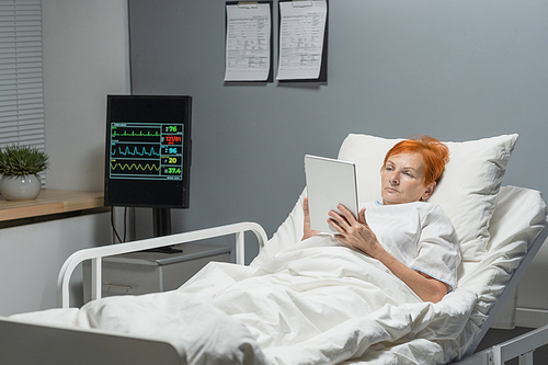 Sick senior woman lying in bed in hospital ward and using digital tablet, she reading online book