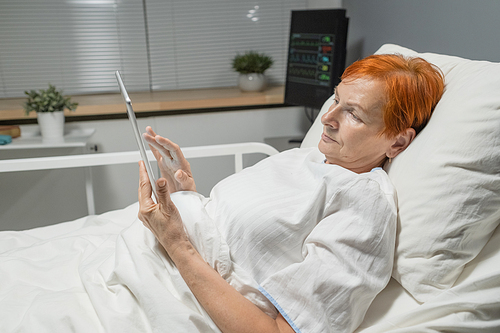 Elderly woman lying in bed and typing on digital tablet, she communicating online while treating at hospital