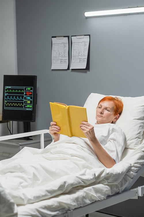 Elderly woman resting in bed for reading an interesting book during her treatment at hospital