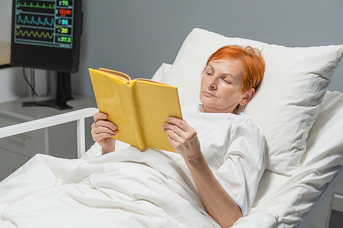 Senior woman lying on bed in hospital ward and reading a book, she recovering after disease