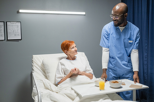Male nurse bringing breakfast for senior woman who lying in bed in hospital ward