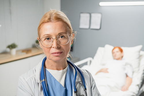 Portrait of mature female doctor in eyeglasses looking at camera while standing at hospital ward with patient in the background