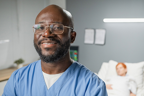 Close-up of African surgeon in eyeglasses smiling at camera while standing at hospital ward with patient in the background